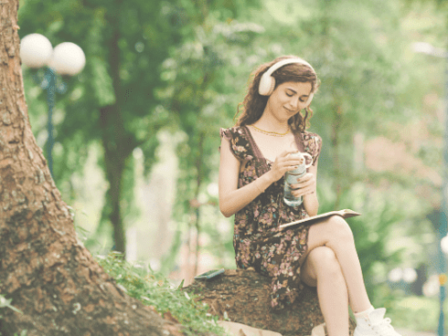Image of a young woman sitting on a tree stump surrounded by bush thoughtfully reading her book while listening to beautiful music through headphones.