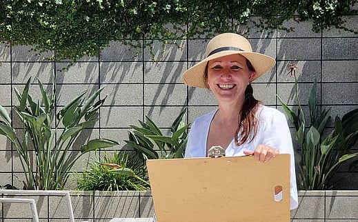 Award winning garden designer, Kerry Speirs, doing an onsite garden design coaching session. Shes's standing in a garden courtyard holding a clipboard and wearing a sun hat. There are plants and outdoor furniture in the background.