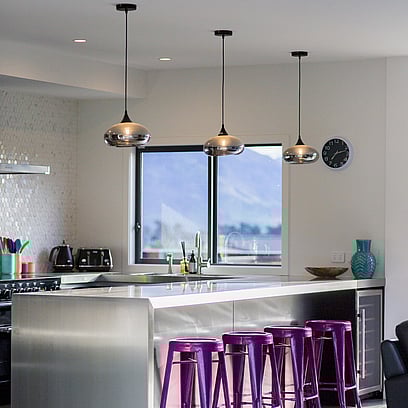 A light and bright kitchen with white bench tops, purple bar stools and feature pendant lights.