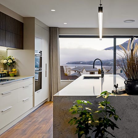 Looking out over a designer kitchen with light stone bench tops, bar pendant lighting, wooden floors and a window view out to the lake.