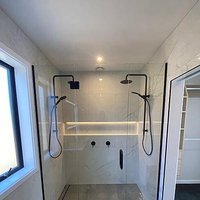 A light and bright white bathroom with large double sized glass shower, black shower hardware, and tiled floor with strip drains.