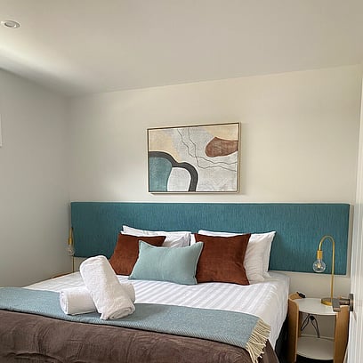 A light walled modern bedroom with an artwork on the wall above a large double bed, teal headboard, throw cushions, and beside table with lamp.