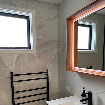 A marble look tiled bathroom with a white topped vanity, black heated towel rail, and framed mirror.