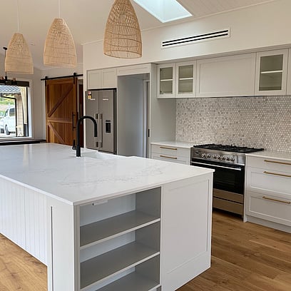 The main house's kitchen featuring timber floors, white bench top and cabinetry, black tap, and woven pendant lights.