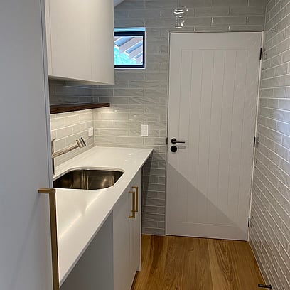 A fully functional laundry with light grey tiled walls, white bench top and white cabinetry with brass handles.