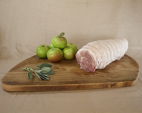 pork shoulder roasting joint on a large wooden chopping board with a pile of bramley apples and a sprig of sage