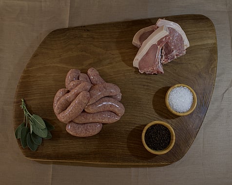 wooden board from above displaying a pile of sausages and three chops with bowls of rock salt and peppercorns & a sprig of sage