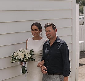 A bride carrying a bouquet of flowers walking along the white wooden south wall of the Kūaotunu Hall, escorted by her father