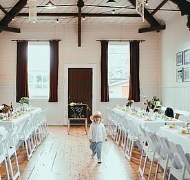 A child wearing a white hat walking between a row of wedding tables and chairs at the Kūaotunu Hall. In the background are two high sash windows with velvet curtains with a red door in between.