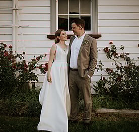 A bride standing with her groom outside the Kūaotunu Hall with a display of Dublin Bay red roses in the garden behind them