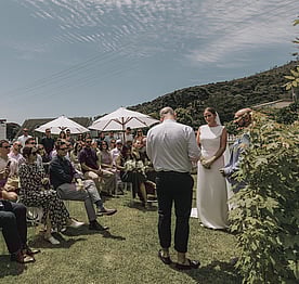 A wedding party on the lawn at the back of the Kūaotunu Hall watching a bride and groom get married