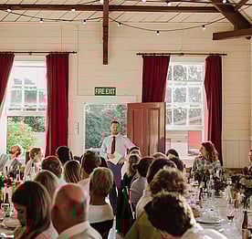 Guests at a wedding at the Kūaotunu Hall listening to a man giving a speech at the far end of the hall, with tall sash windows flanked by red velvet curtains on either side of him and the hall's red front door behind him