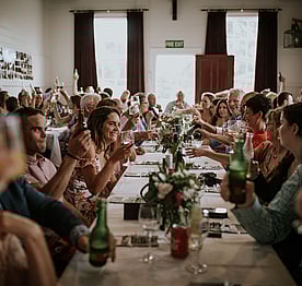 Wedding guests sitting at rows of tables in the Kūaotunu Hall raising their glasses for a toast