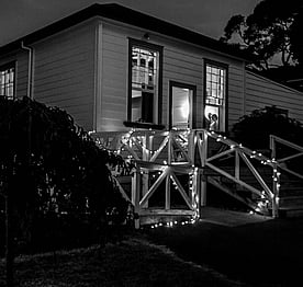 A black and white photo showing the exterior of the Kūaotunu Hall building, including the ramp, lit up for a wedding