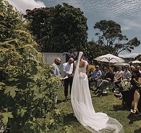 A bride and groom standing on the back lawn of the Kūaotunu Hall surrounded by wedding guests watching the ceremony