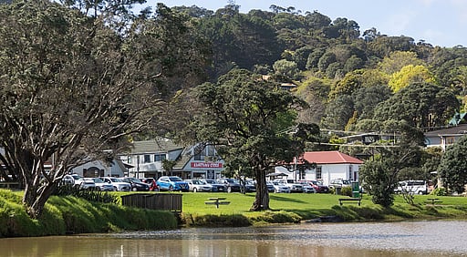 A view of the Kūaotunu Hall as seen from the other side of the Kūaotunu river. The white painted wooden hall with its red roof can be seen in the distance next to the Kūaotunu store and a row of cars parked by the riverside.
