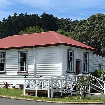 The Kūaotunu Hall as seen from the front - a white painted wooden building with a red roof and red door, with steps leading up to it and a ramp to the left