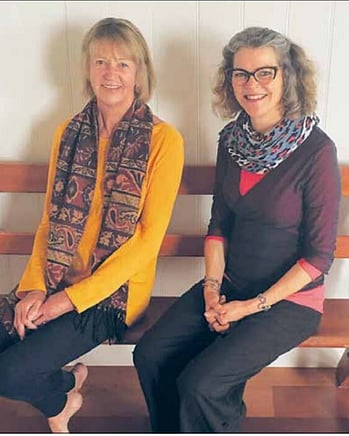 Kūaotunu Hall Committee member Jo Mullins (left) sitting with yoga teacher Jose Kakebeeke (right) on one of the kauri pews in the Kūaotunu hall