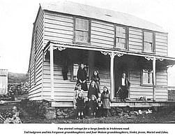 An historic black and white photo showing a two storied wooden building in Irishtown Rd in Kūaotunu with a man with a pipe in his mouth sitting on the front verandah and his 7 granddaughters standing to the left of him