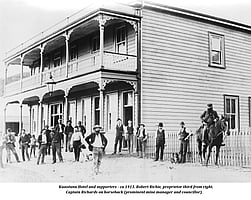 An historic black and white photo of the two-storied wooden Kūaotunu hotel, circa 1913, with several patrons standing outside and a man on horseback
