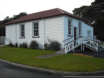 The exterior of the Kūaotunu Hall, a white-painted wooden building with a red roof and with a ramp plus steps up to the front door