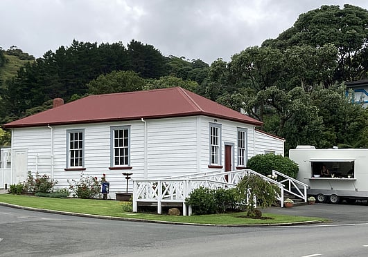 The exterior of the historic Kūaotunu Hall, with its white wooden walls and a red roof, and steps leading up to the front door plus a ramp to the left