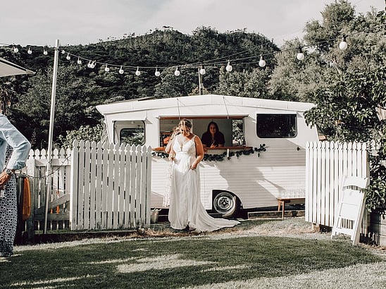 A bride with a glass of champagne standing outside a caravan bar parked by the white picket fence outside the Kūaotunu Hall