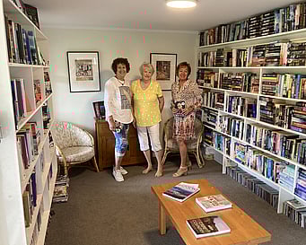 The 3 women members of the inaugural Kūaotunu Library Committee standing inside the library with rows of bookshelves on each side of them