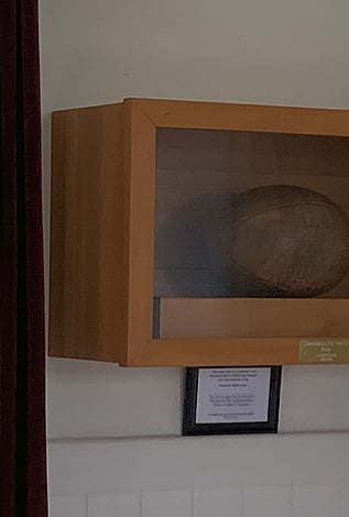 A rugby ball displayed in a wooden case, commemorating a century of rugby in Kūaotunu