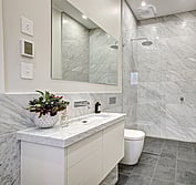 Interior view of ensuite showing stone vanity, wall hung toilet and open shower with glass screen. Ensuite with honed Carrara marble wall tiles and vanity with cut in sink. Honed black granite floor tiles.