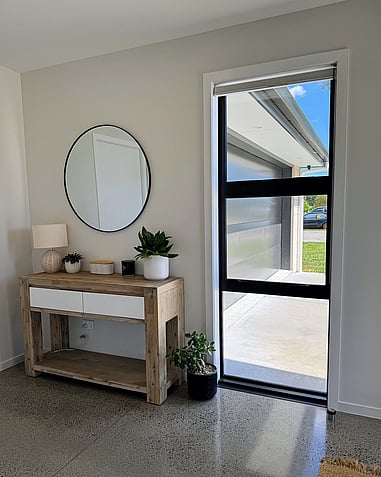 Image of front entry of new home. Sunlit with tiled flooring. Styled with light timber credenza and circle mirror. Home built by Coull Builders, Otorohanga.