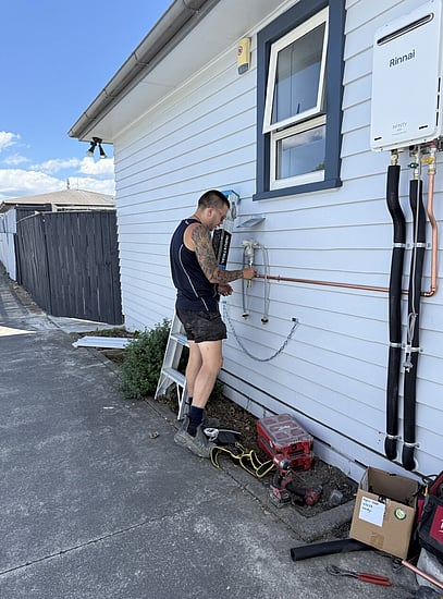 plumber installing rinnai inifinity gas hot water system on the side of a white weatherboard single story house