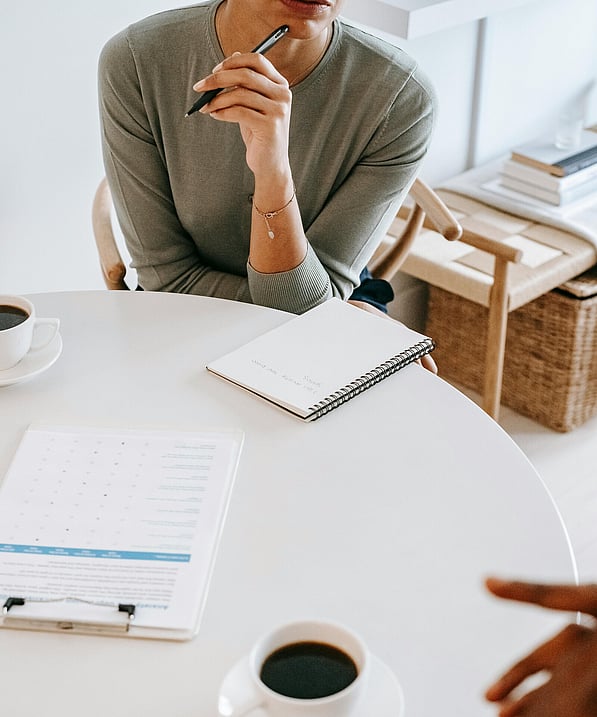 woman sitting on a round table, having a conversation with a client. She has a notepad in front of her, a clipboard and two cups of coffee are showing.