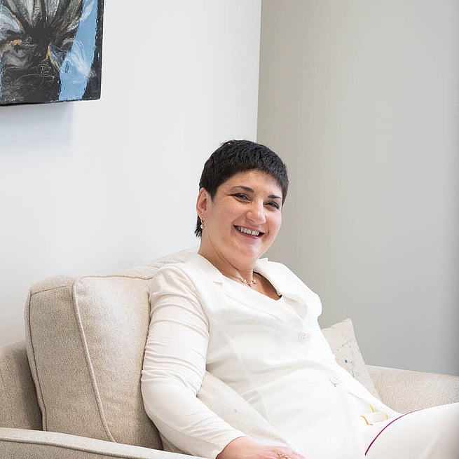 Smiling woman with short dark hair wearing a white outfit, seated in a beige armchair in a bright office space.