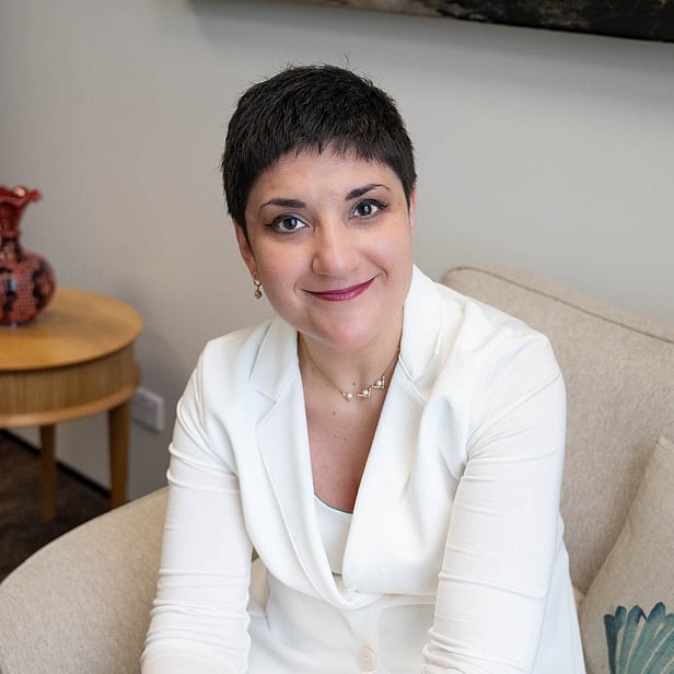 Smiling woman with short dark hair wearing a white jacket, seated on a beige sofa in an office setting.