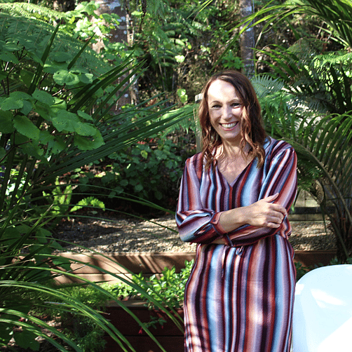 A woman wearing a blue and red striped dress striped dress. She has shouder length brown hair. Her arms are crossed and shes smiling.