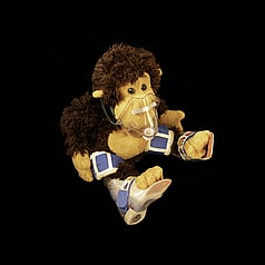 Soft toy monkey wearing AFOs and surgical gas mask. It is centrally placed on a square black background.