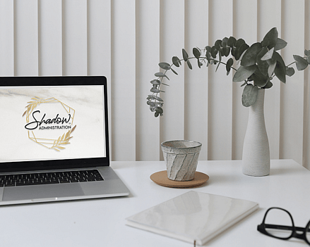 Laptop open on a virtual assistants desk with the Shadow Administration logo on the screen. Notebook and glasses on a white desk.