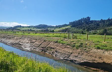 Rural landscape with grassy paddocks, fence line and narrow stream running through farmland under clear blue sky