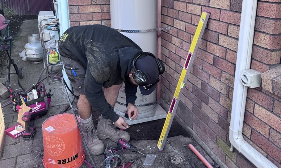 plumber wearing black sweatshirt leaning over fitting a new exterior hot water cylinder beside a red brick exterior house wall