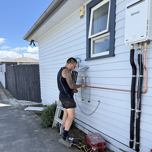 plumber weating black singlet working outside white weatherboard house fitting a gas infinity hot water system
