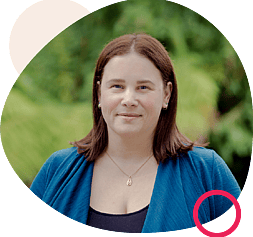 Smiling woman with shoulder-length brown hair, wearing a blue cardigan and pendant necklace, framed by abstract beige and pink shapes