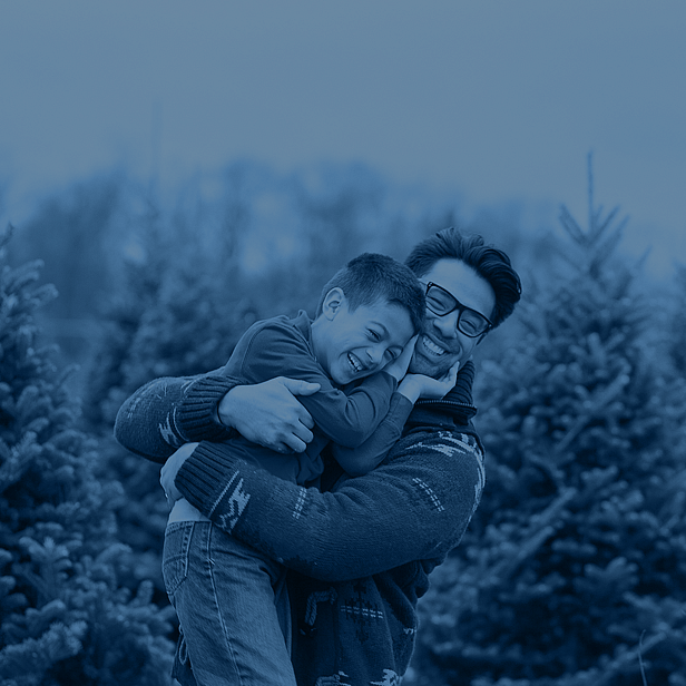 A father holding his young son while they laugh together outdoors in a wintery setting, surrounded by pine trees, both dressed warmly for the cold weather