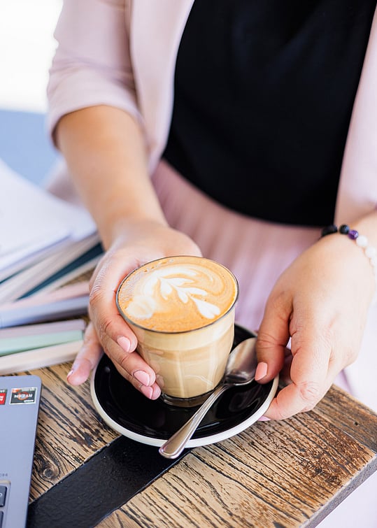 A woman holding a latte in a glass cup with latte art, seated at a wooden table with notebooks and a laptop - capturing a calm, focused moment during a CV consultation session.
