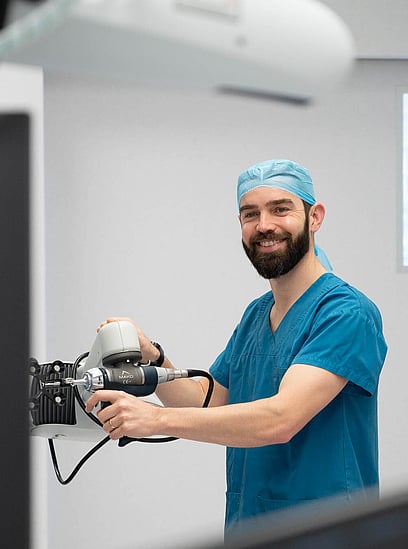 Hamish McLaren, a surgeon in blue scrubs, smiling while operating a robotic surgical device in a medical facility.