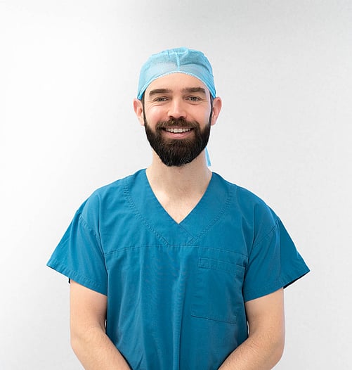 Hamish McLaren n an operating room, wearing surgical scrubs and a cap, operating a robotic surgical device while another medical professional works in the background.