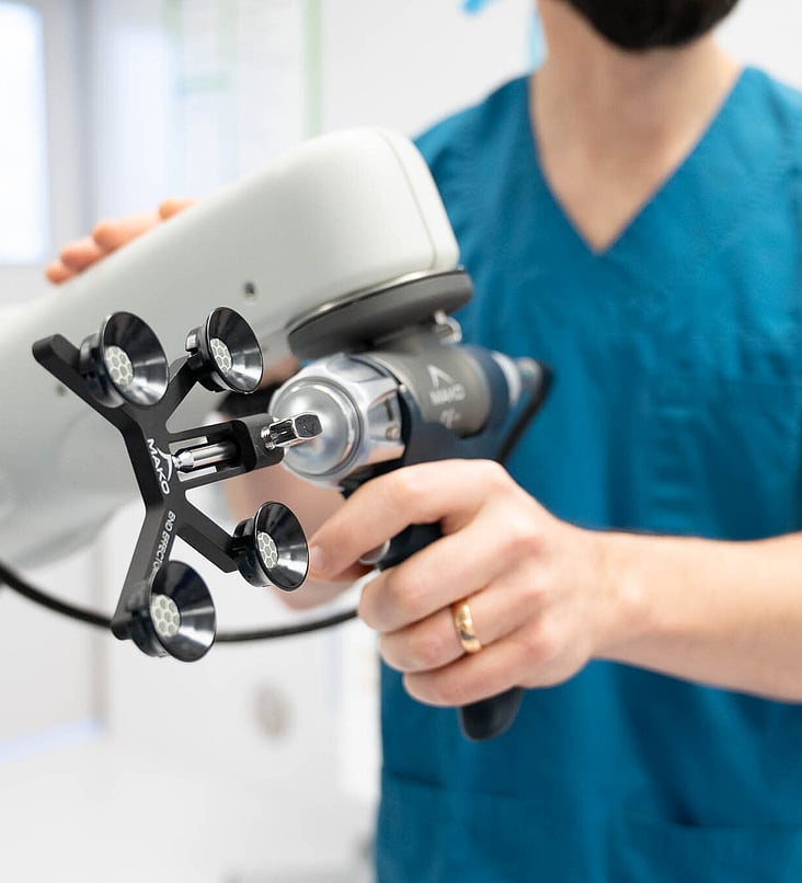 Hamish McLaren n an operating room, wearing surgical scrubs and a cap, operating a robotic surgical device while another medical professional works in the background.