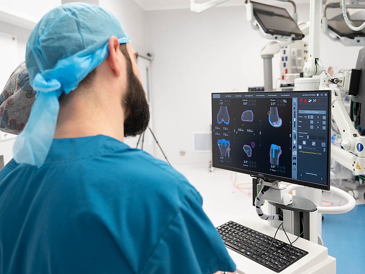Hamish McLaren n an operating room, wearing surgical scrubs and a cap, operating a robotic surgical device while another medical professional works in the background.