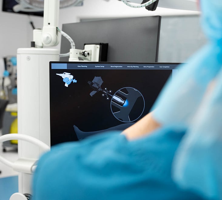 Hamish McLaren n an operating room, wearing surgical scrubs and a cap, operating a robotic surgical device while another medical professional works in the background.