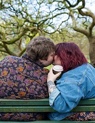 Couple share a kiss, coffee in hand during their walk through a beautiful park